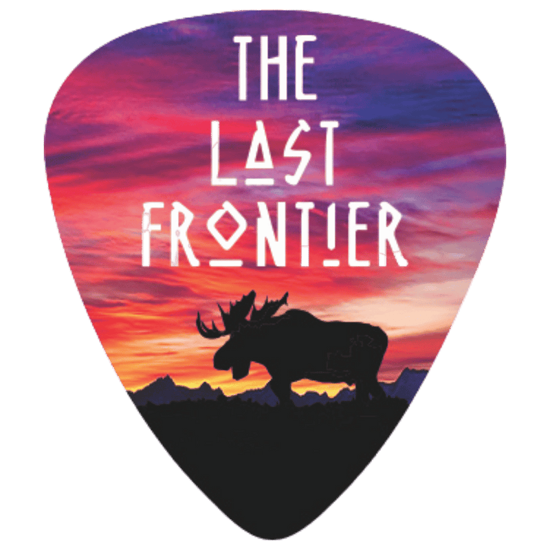 A guitar pick featuring a colorful sunset landscape design with the text 'THE LAST FRONTIER' and a black moose silhouette over mountains.