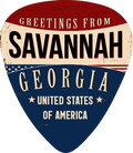 A guitar-pick-shaped sign or sticker with a distressed patriotic design reading 'Greetings from Savannah, Georgia, United States of America' in cream lettering on red, beige, and dark blue sections.